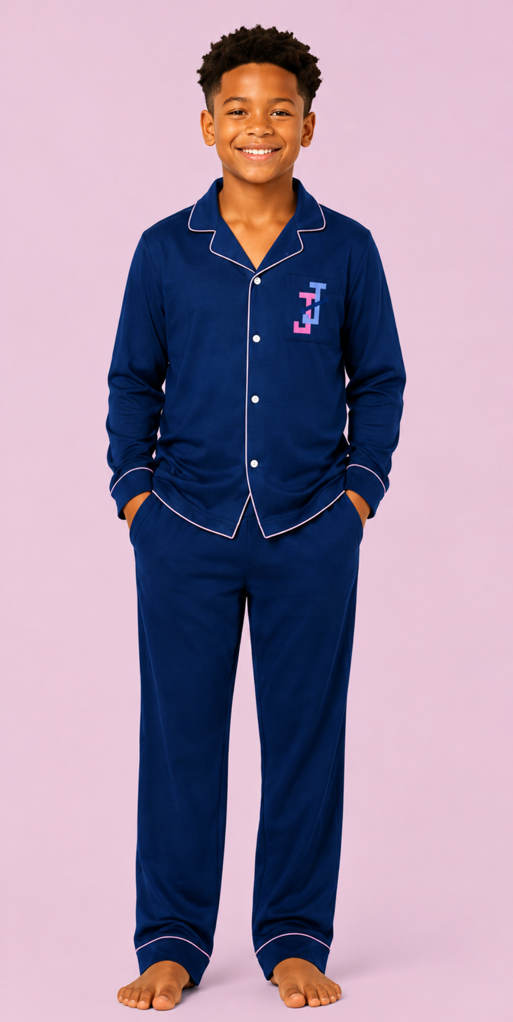 A young boy wearing a navy blue pajama set with white piping and a small embroidered monogram on the chest, standing against a solid light pink background.
