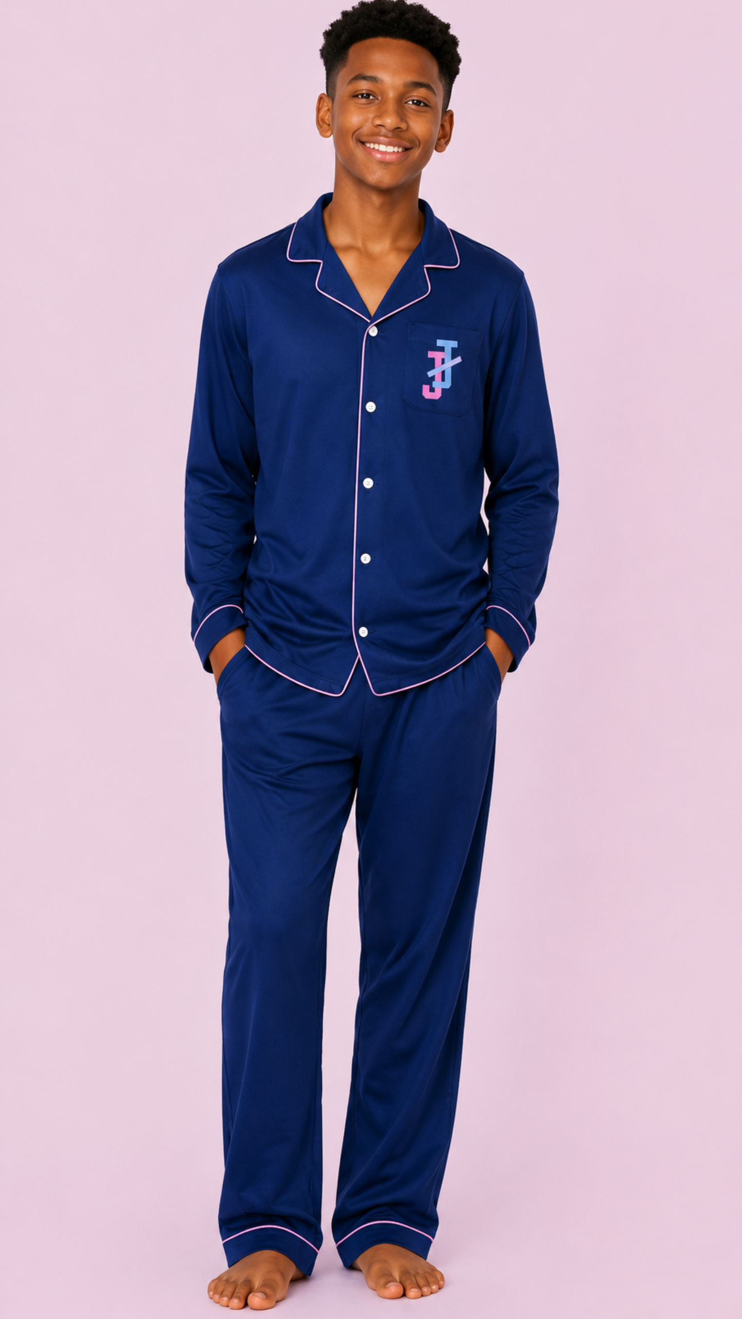 A teenage boy wearing a navy blue pajama set with white piping and a pink embroidered monogram logo on the chest, standing against a solid pink background.