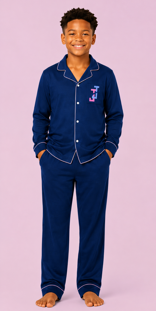 A young boy wearing a navy blue pajama set with white piping and a small embroidered monogram on the chest, standing against a solid light pink background.