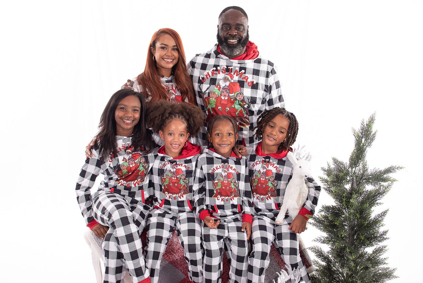 Family of six wearing matching black and white checkered pajamas with red accents, standing in front of a small Christmas tree on a white background.